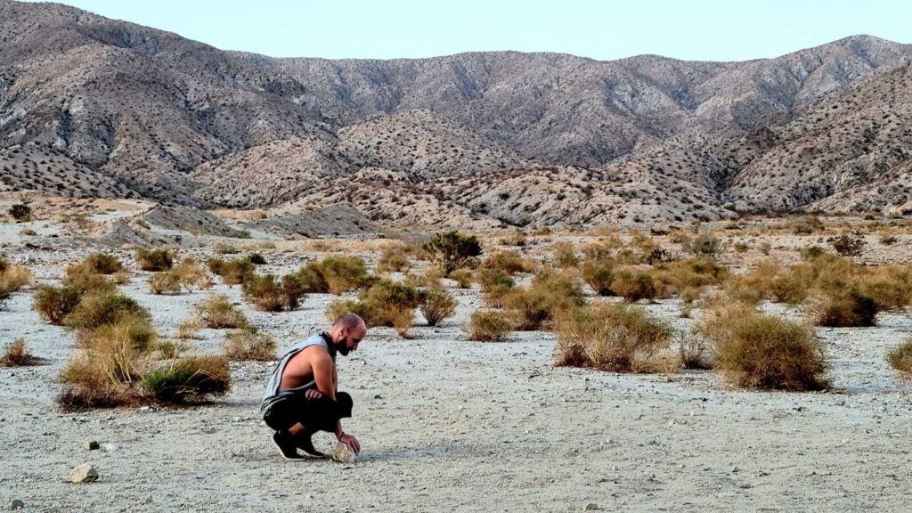 Scholarly Communications Lab member Mario Malicki crouches down to touch a rock on the dusty desert floor of Desert Hot Springs, California. In the background: endless shrubs and mountains