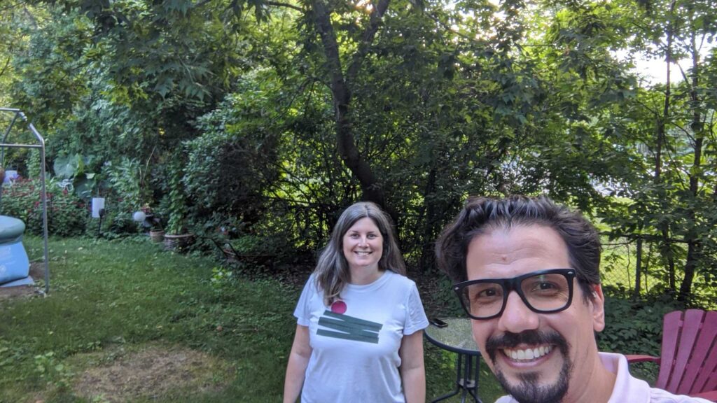 ScholCommLab co-directors Stefanie Haustein and Juan Pablo Alperin smile for the camera against a sunlit backdrop of trees. 