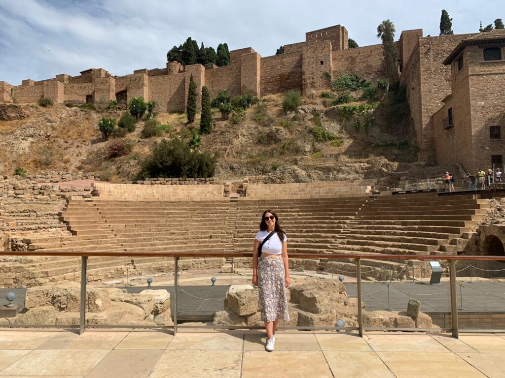 Maddie Hare, member of the Scholarly Communications Lab, smiling into the camera in front of Roman Theatre at the Alcazaba in Málaga, Spain